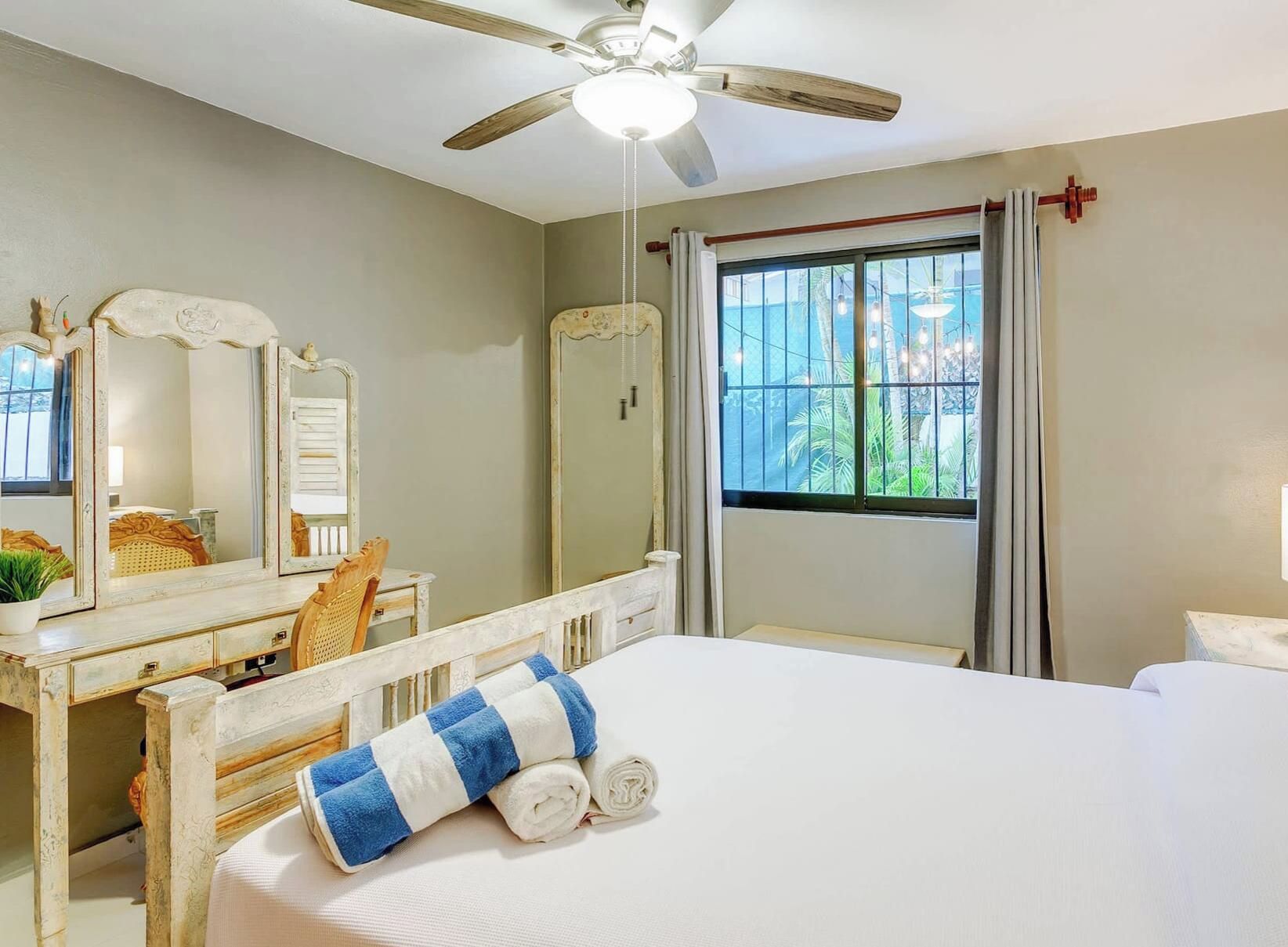 Quarto com cama, escrivaninha, cadeira, ventoinha no teto e janela com cortinas.