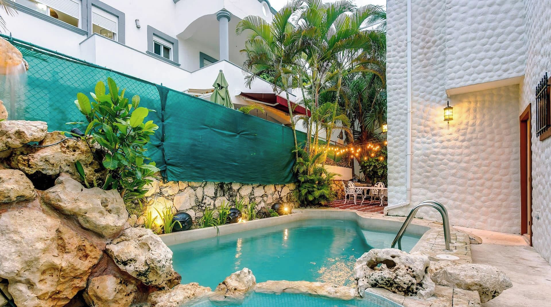Une piscine bordée d’un mur en pierre et entourée de plantes tropicales, le tout adossé à un bâtiment blanc doté d’un balcon.