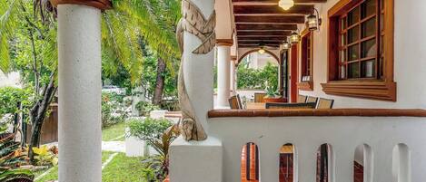 A covered outdoor area with a wooden ceiling, a seating area with a table and chairs, and a view of a garden with various plants and a palm tree.