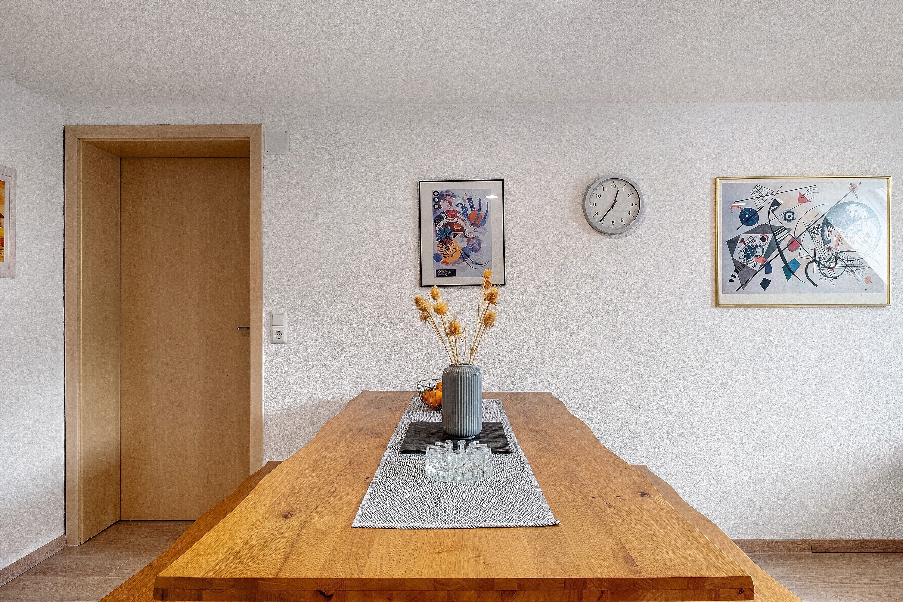 Une table de salle à manger en bois, ornée d’un vase de fleurs séchées, de deux œuvres d’art abstraites encadrées et d’une horloge murale.