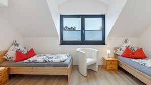 A bedroom with a sloped ceiling, a window, a bed with floral bedding, a white chair, and a bedside table with a lamp. - Deluxe Apartment Lea with Balcony and Wi-Fi (Radolfzell am Bodensee)