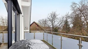 Ein überdachtes Auto auf einem Balkon mit Metallgeländer, ein Gebäude mit großen Fenstern und kahle Bäume im Hintergrund.