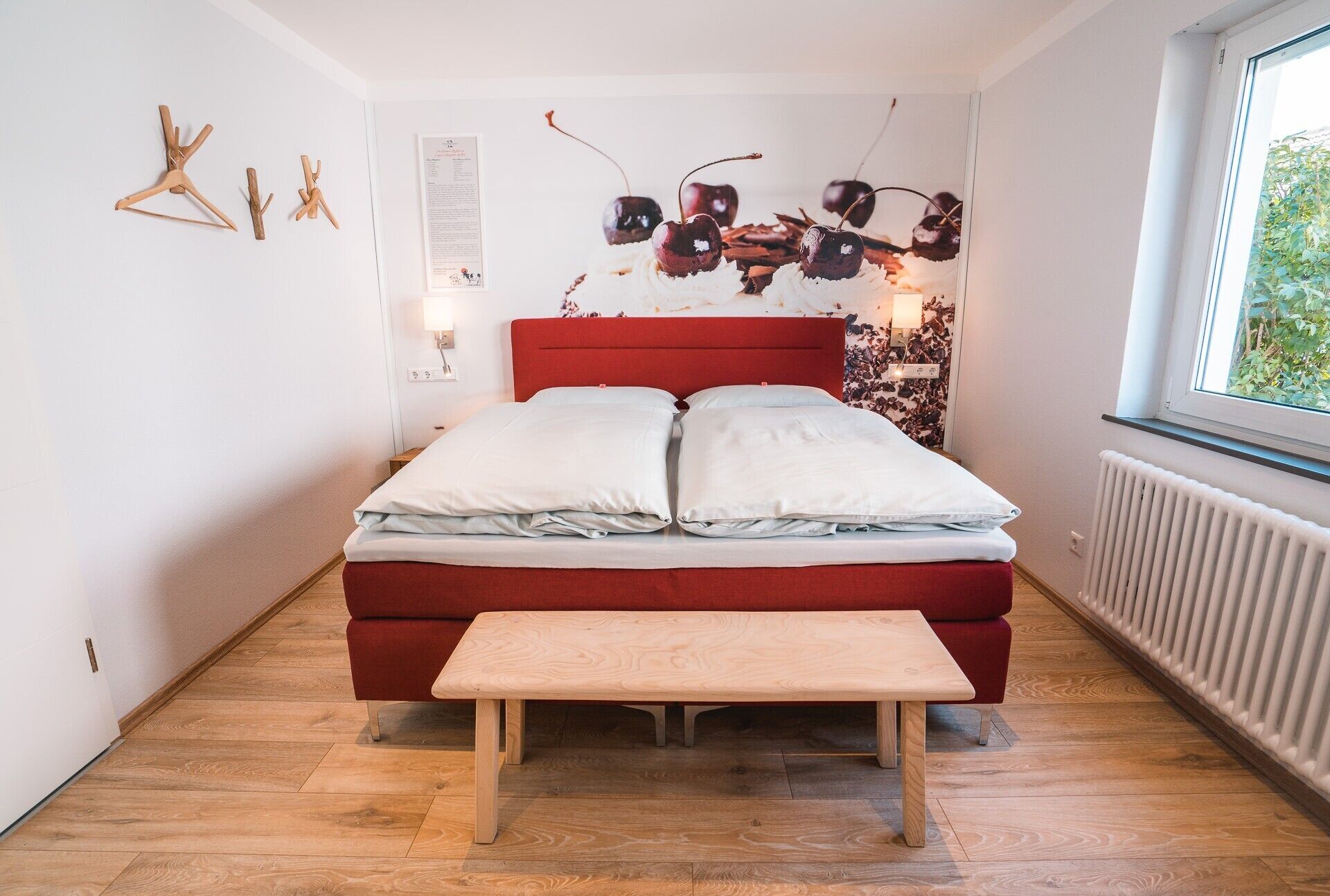 A bedroom with a red bed, white pillows, a wooden bench, a radiator, and a window with a view of greenery.