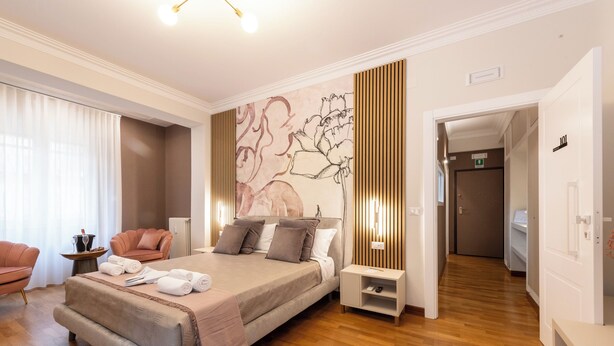 A modern hotel room with a large bed, a sitting area with a chair, a small table with a drink and glasses, and a wall with a floral mural. - Querini Suite (Rome)