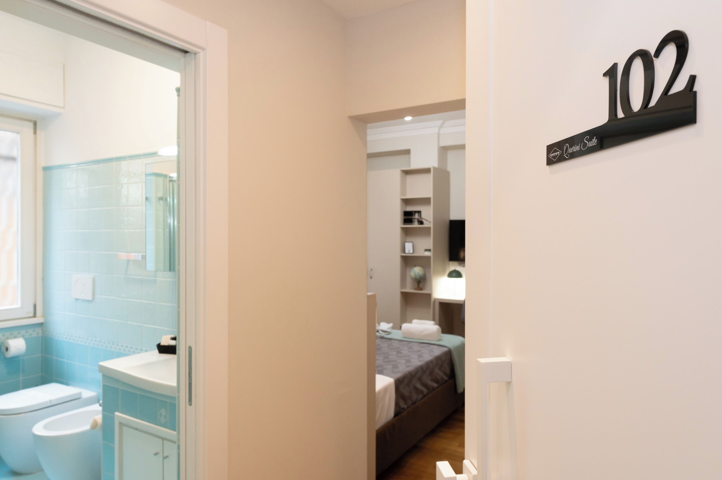 A bathroom with a toilet, sink, and blue-tiled walls. A bed with a gray blanket is visible in the adjacent room.