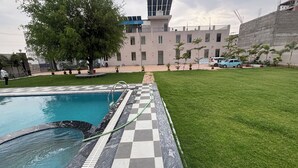 A swimming pool with a checkered border, a grassy area, and a building with a solar panel in the background. - Shree Jagannath Resort (Shri Madhopur)