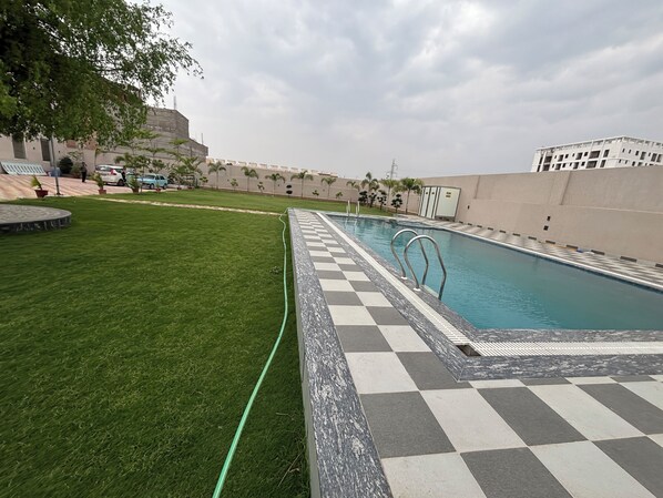 A swimming pool with a checkered border, surrounded by a well-maintained lawn and a few potted plants. - Shree Jagannath Resort (Shri Madhopur)
