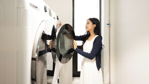 A person operating a front-loading washing machine in a room with a door and a pipe on the wall. - Loft13 Hostel (Seoul)