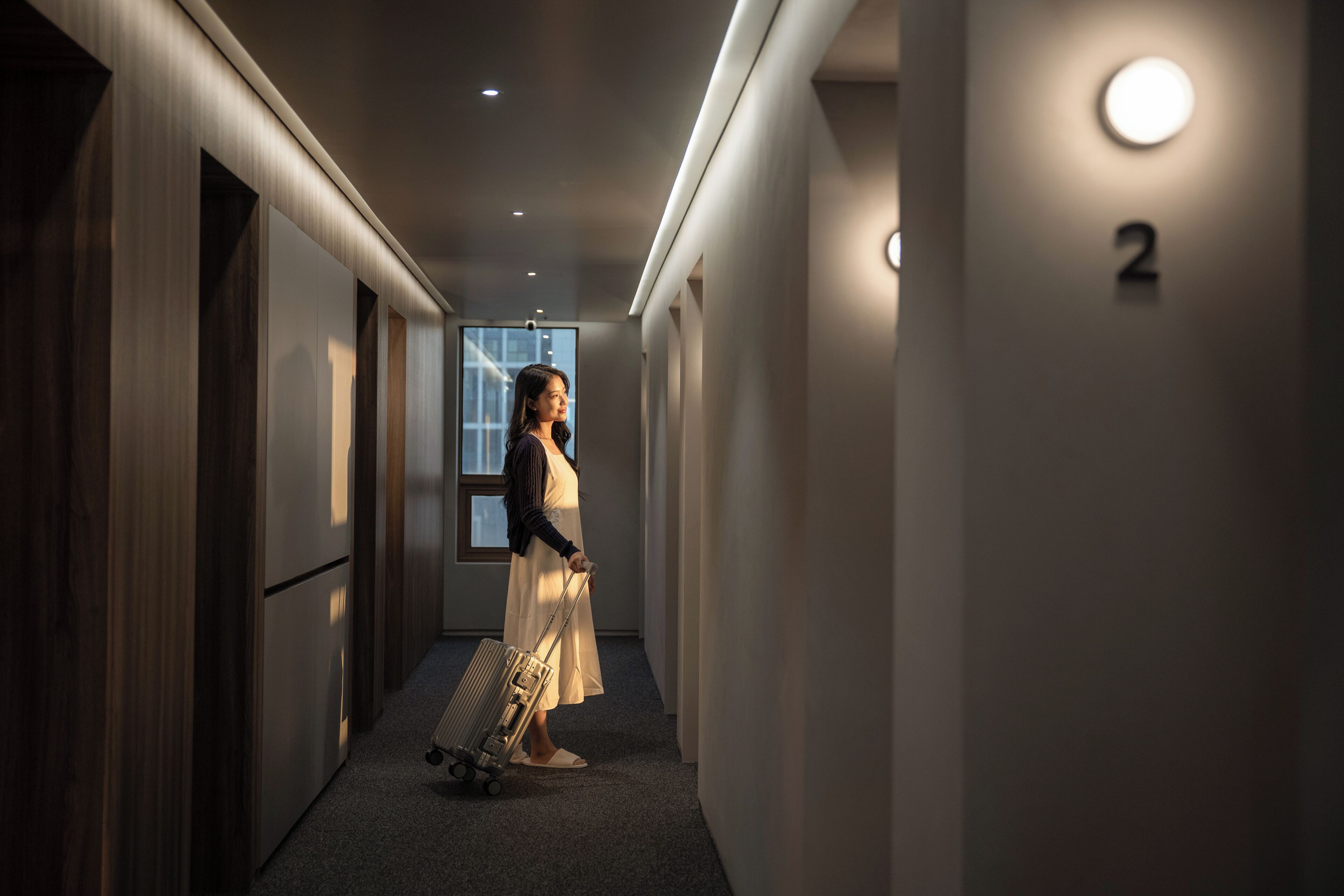 A hotel hallway with a person pulling a suitcase, a number '2' sign, and a window at the end.