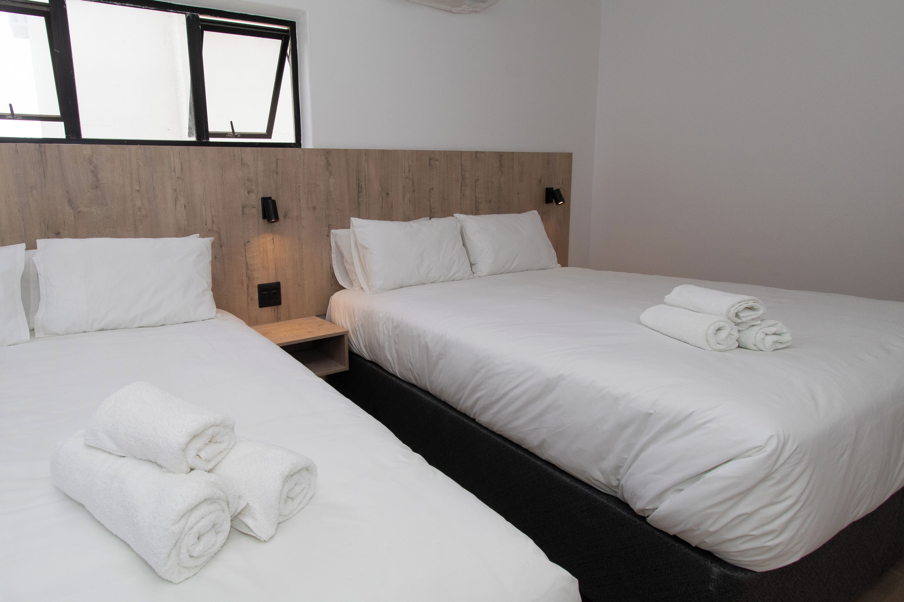 A hotel room with two beds, each with white linens and rolled towels. There is a wooden headboard and a nightstand with a lamp.