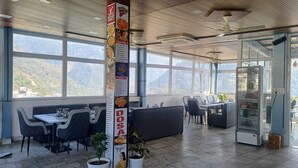 A restaurant with a high ceiling, large windows, and a menu board. There are tables and chairs for dining, and a counter with a display case. - The Hotel JRS (Rishikesh)