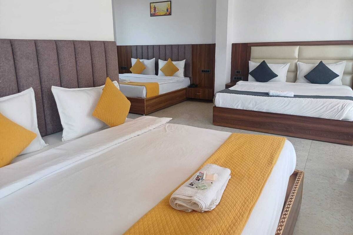 Habitación de hotel con dos camas, cada una con ropa blanca y almohadas amarillas. Una cama tiene cabecera de madera y la otra, cabecera color beige. Una toalla doblada está sobre la cama con la cabecera beige.