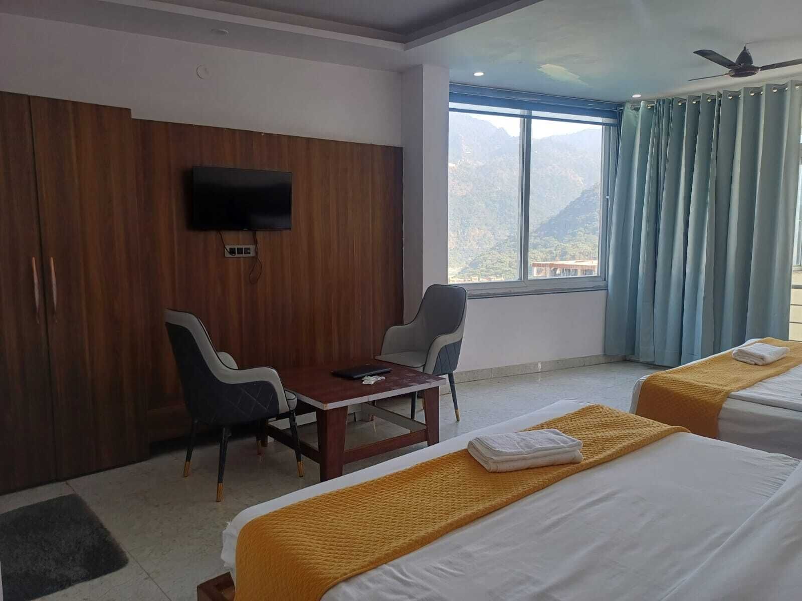 A hotel room with two beds, a desk, a chair, a TV mounted on the wall, and a large window with a view of mountains.