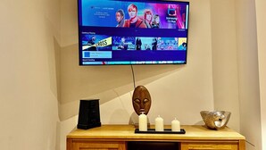 Ein an der Wand befestigter Fernseher zeigt die Benutzeroberfläche eines Streaming-Dienstes, darunter befindet sich eine Holzkonsole mit einer dekorativen Maske, Kerzen und einer Schale.