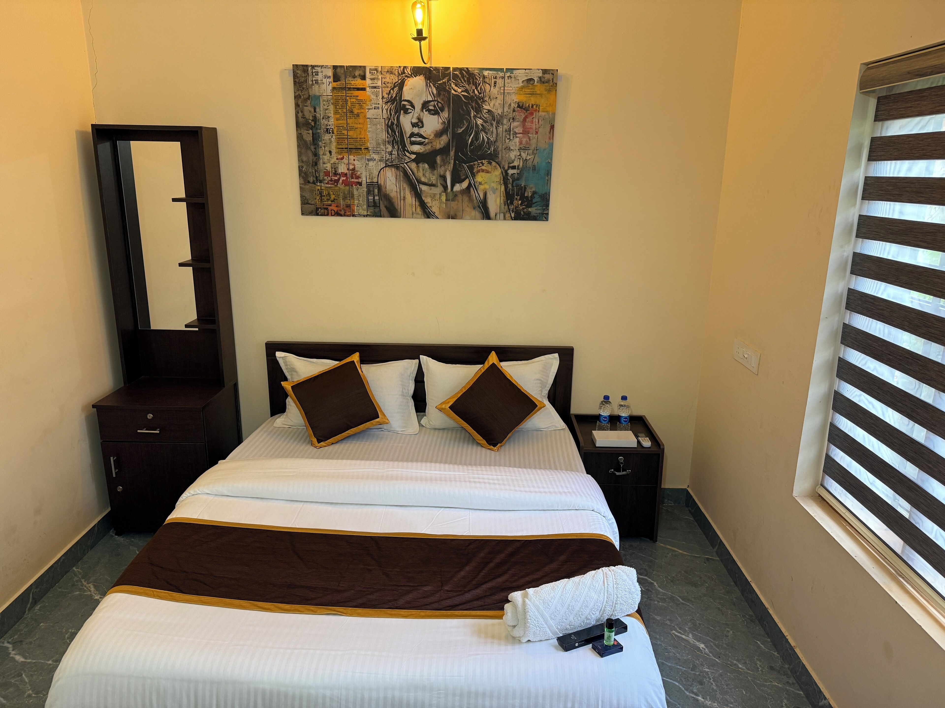 A hotel room with a bed, a nightstand, a mirror, a painting, and a window with blinds.
