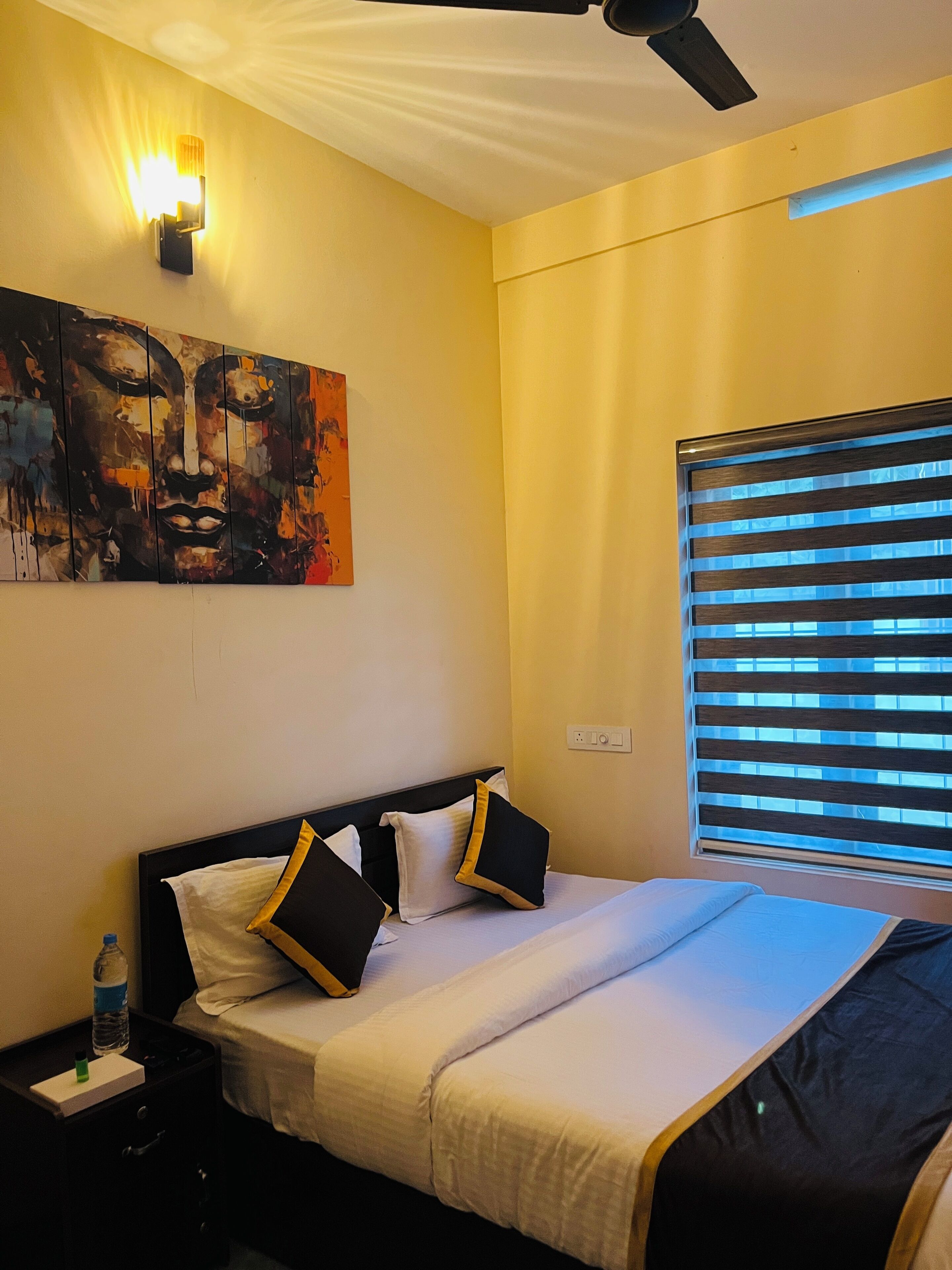 A bedroom with a bed, a nightstand, a wall-mounted light, a ceiling fan, and a window with blinds.