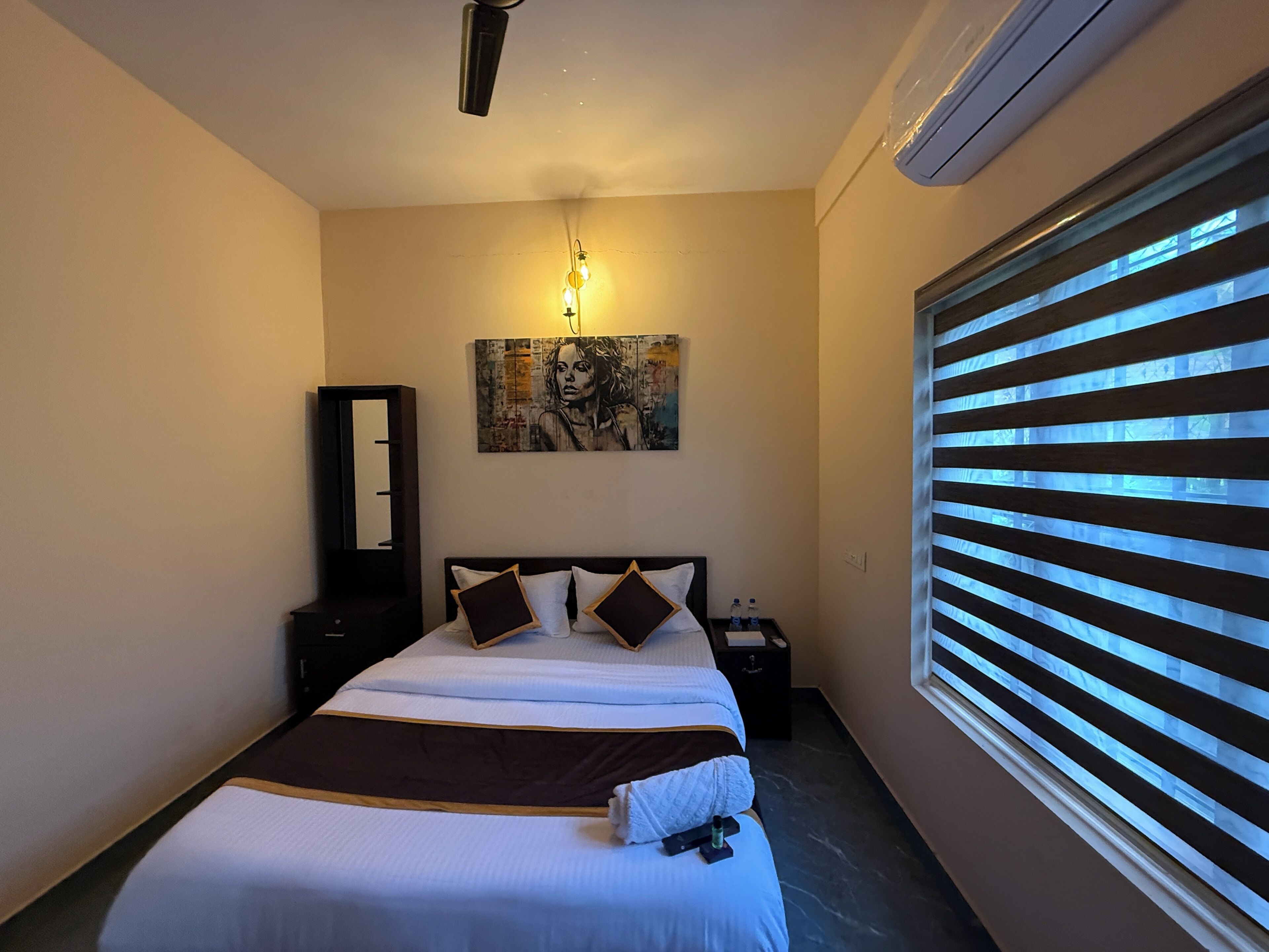 A hotel room with a bed, a bedside table, a mirror, a painting on the wall, and a window with blinds.