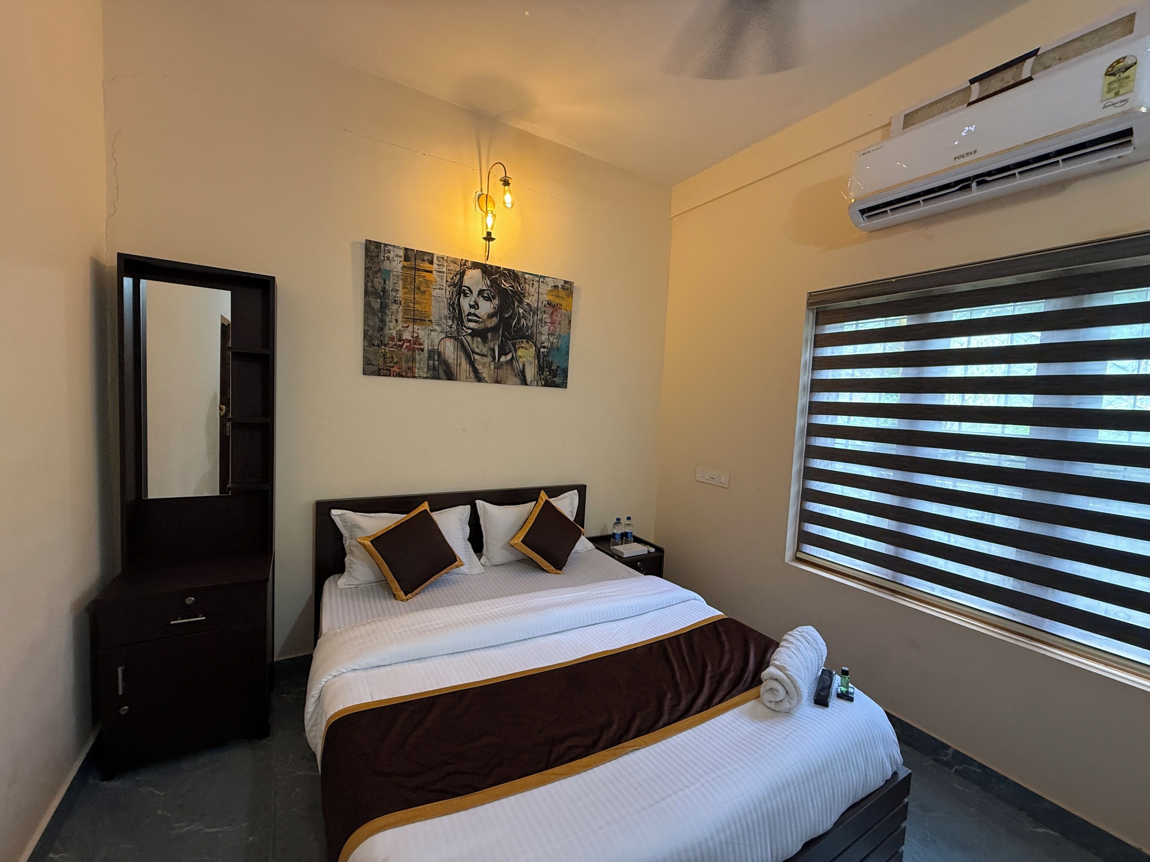 A bedroom with a bed, a nightstand, a mirror, an air conditioner, and a window with blinds.