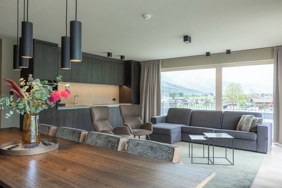 Ein modernes Wohnzimmer mit Essbereich, grauen Sofas und einem großen Fenster mit Blick ins Freie.