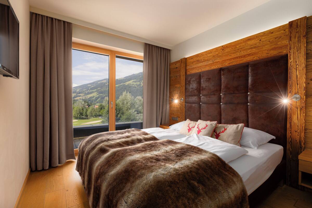 Ein Schlafzimmer mit einem großen Bett, einem hölzernen Kopfteil, einem an der Wand befestigten Fernseher und Blick auf eine gebirgige Landschaft durch ein großes Fenster.