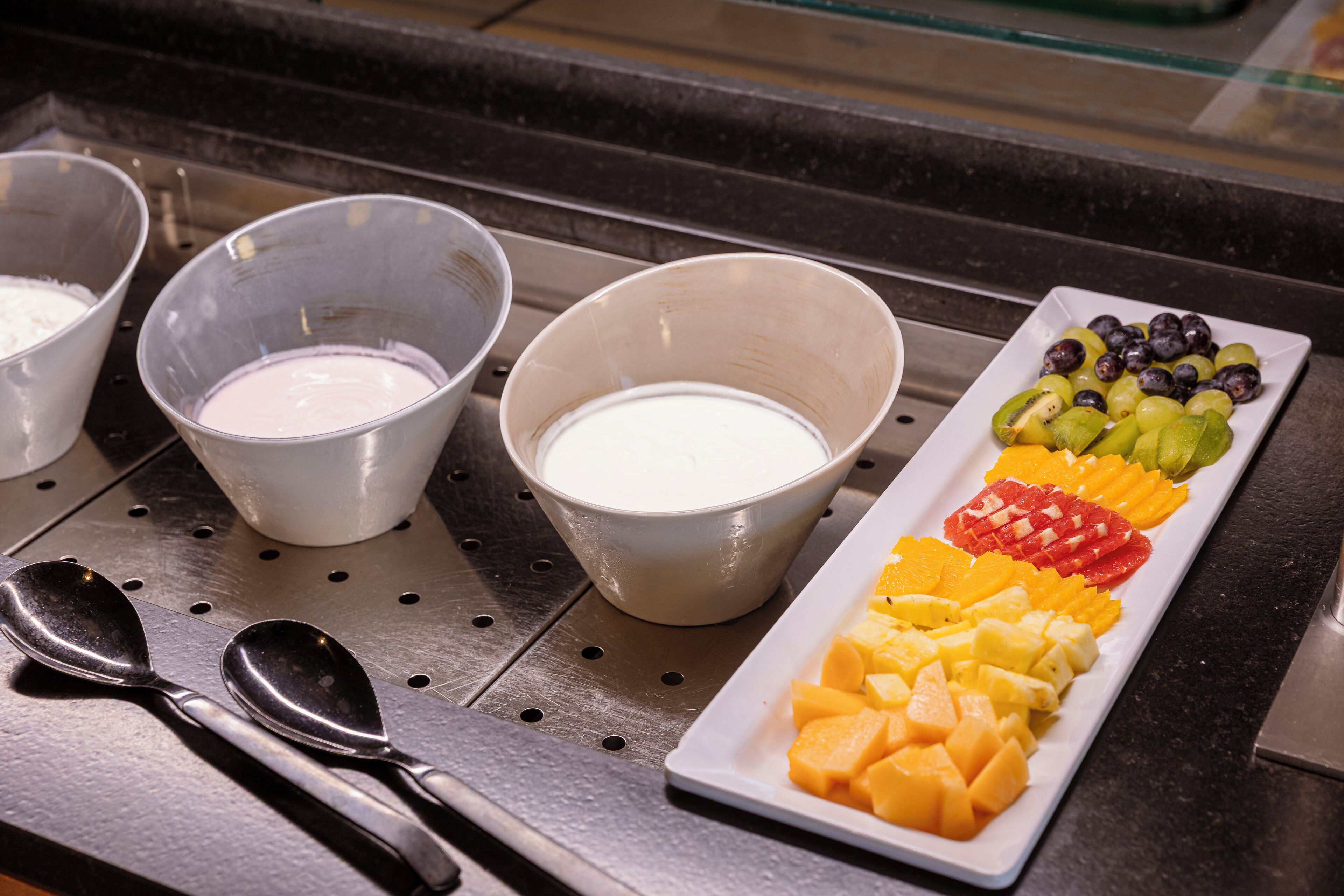 Un buffet avec des bols de sauces blanches et roses, une assiette de fruits variés et deux cuillères posées sur un comptoir noir.