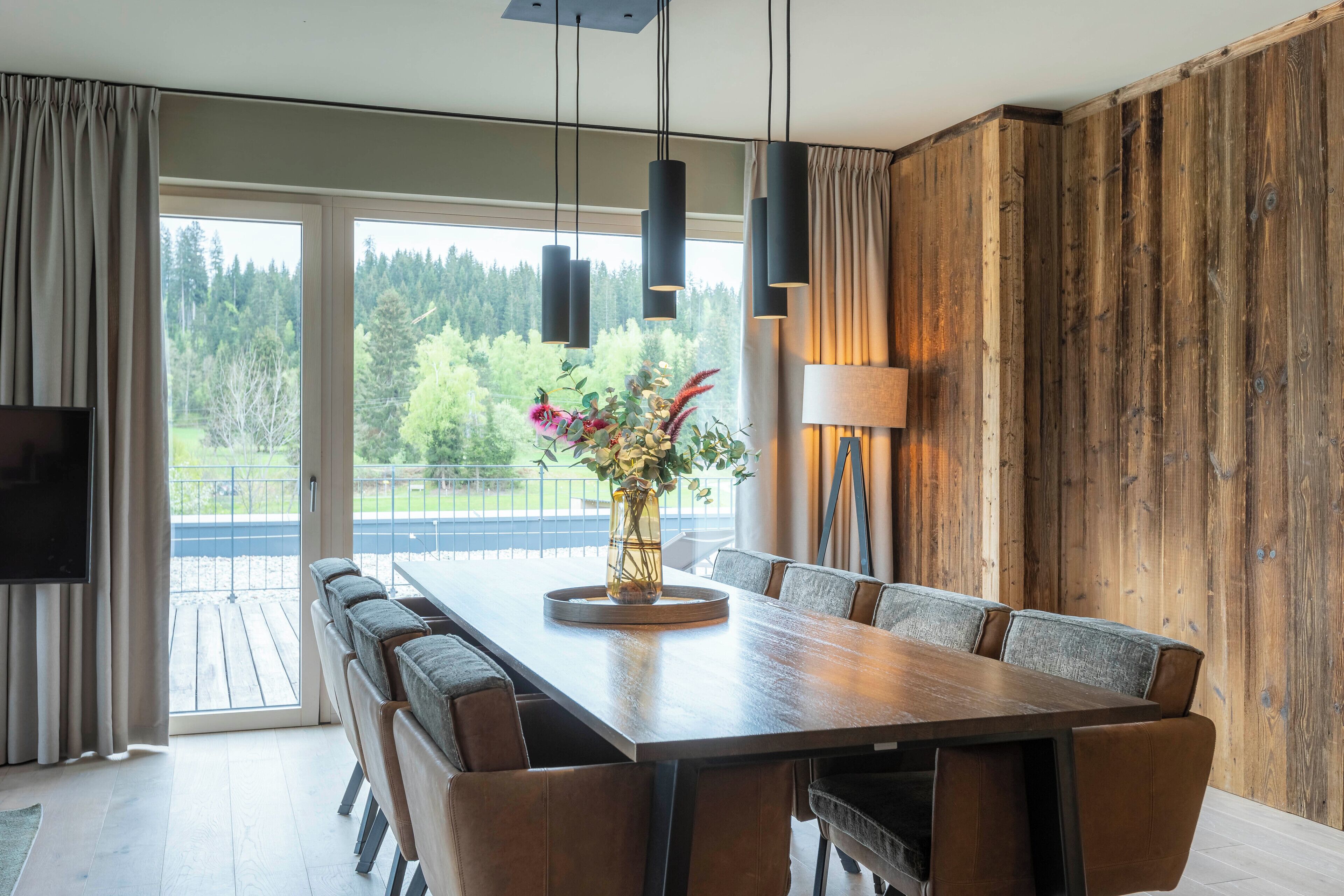A dining area with a long table, multiple chairs, a vase with flowers, hanging lights, and a view of greenery outside.