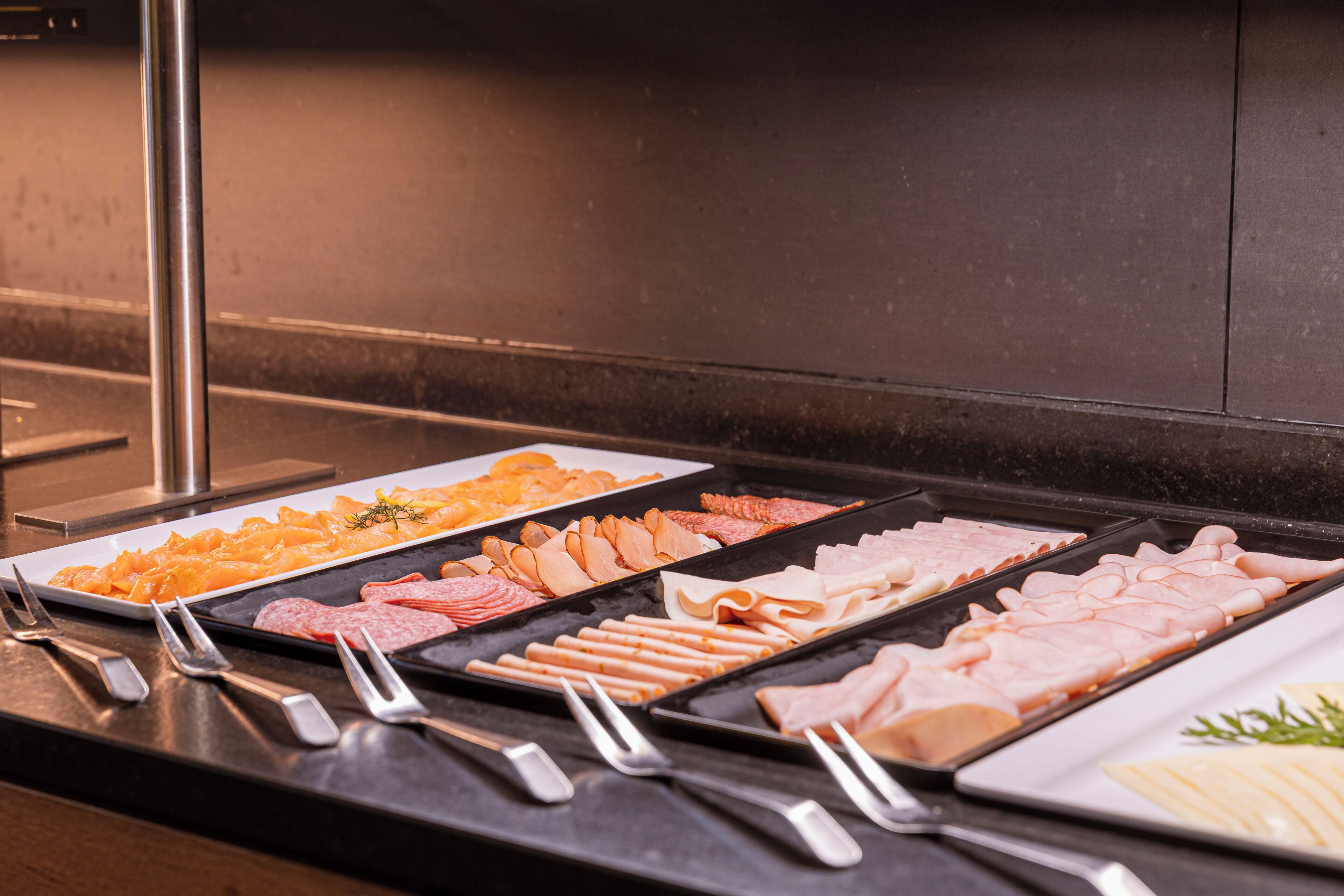 A buffet with various meats and cheeses, accompanied by serving utensils.