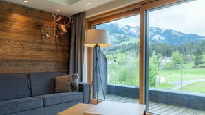 A modern living room with a grey sofa, a wooden coffee table, and a floor lamp. A large window offers a view of a green landscape and mountains. - Berg & Golf Apartments Westendorf by ALPS RESORTS (Westendorf)