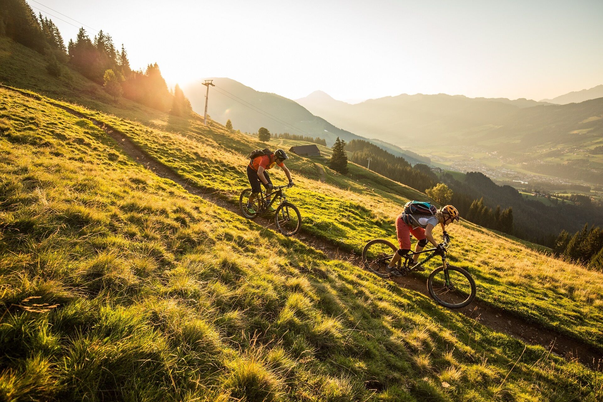Dos ciclistas en un sendero montañoso cubierto de hierba, con una vista panorámica de colinas lejanas y un pequeño edificio al fondo.