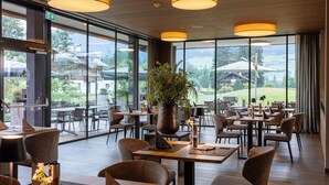 A spacious indoor dining area with large windows, modern furniture, and a central plant. - Berg & Golf Apartments Westendorf by ALPS RESORTS (Westendorf)