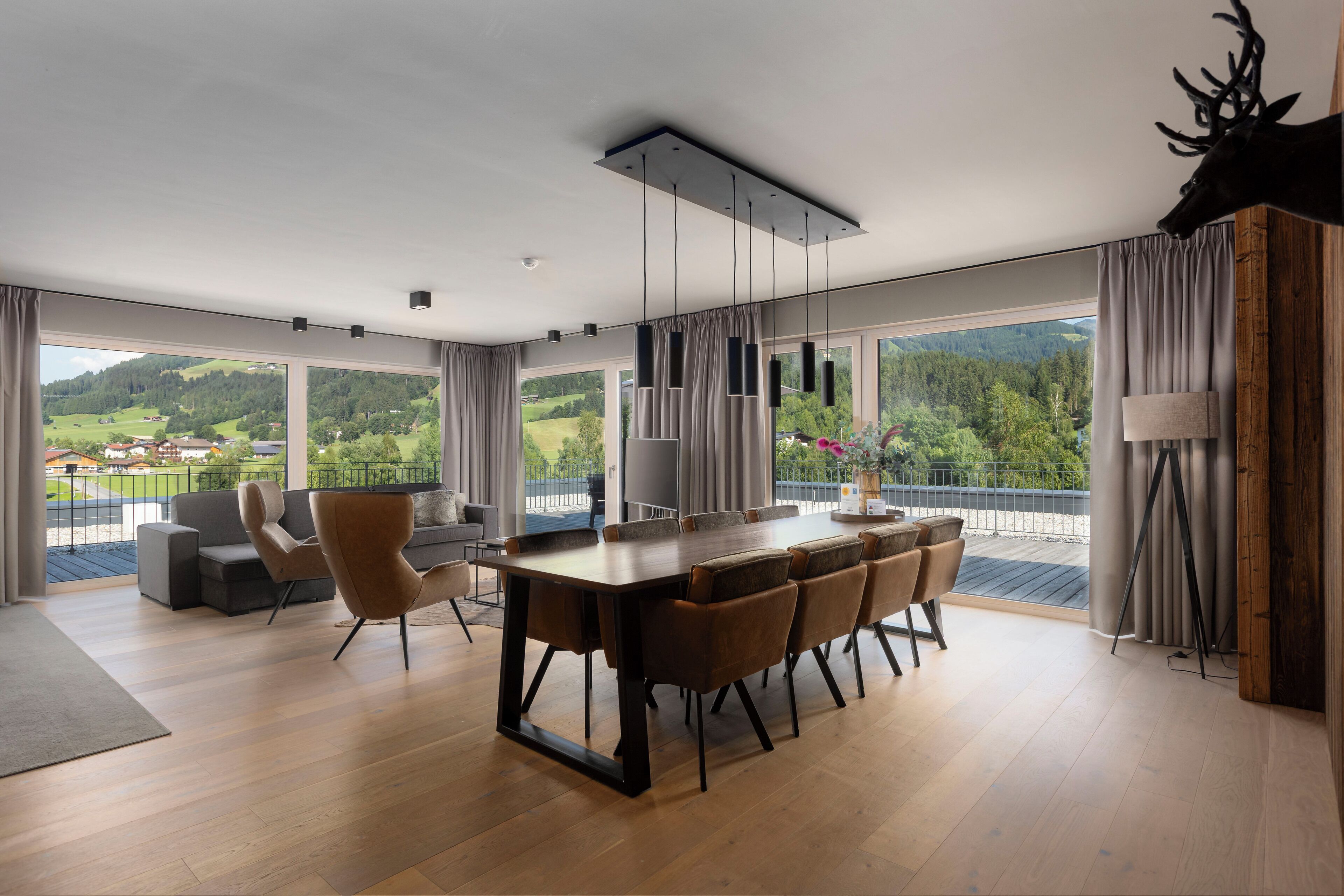 A modern dining area with a wooden table and chairs, large windows with curtains, and a view of a green landscape.