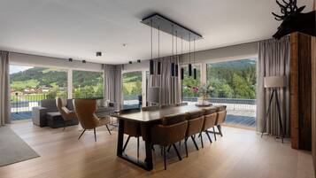 A modern dining area with a wooden table and chairs, large windows with curtains, and a view of a green landscape.