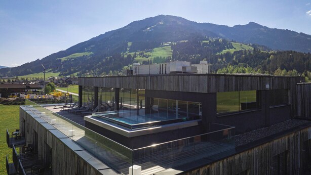A modern building with a glass-enclosed pool area and a view of a mountain range. - Berg & Golf Apartments Westendorf by ALPS RESORTS (Westendorf)