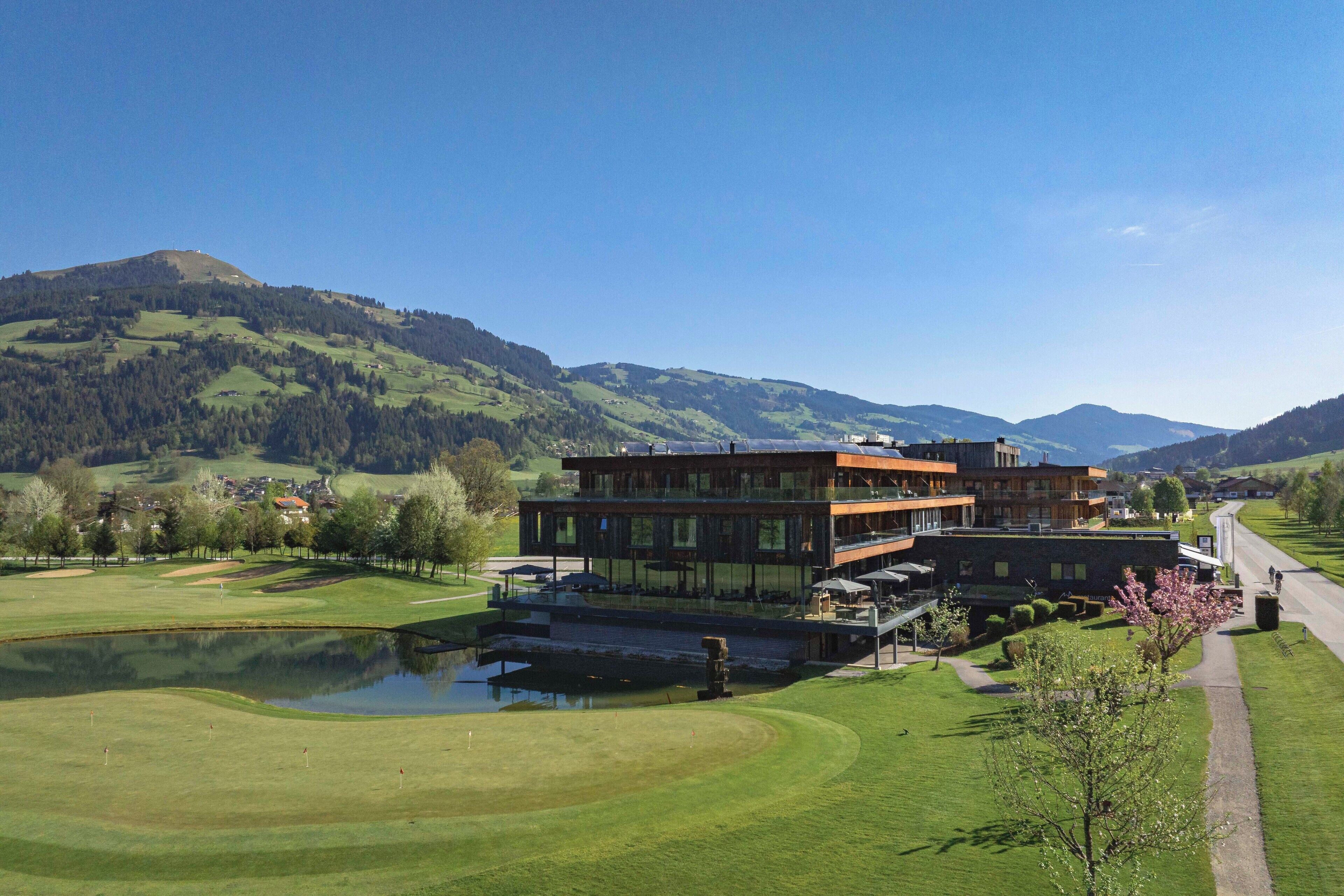 A modern building with a glass facade, situated on a golf course with a pond and a clear view of the surrounding hills.