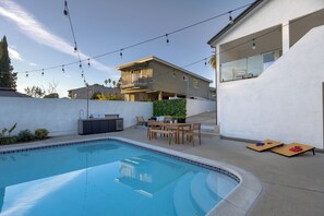 Ein moderner Poolbereich im Freien mit Essplatz, Lichterketten und einem zweistöckigen Haus im Hintergrund.