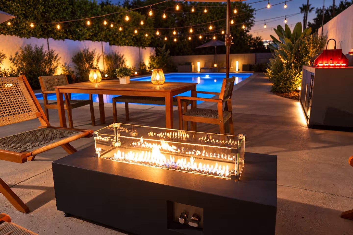 An outdoor patio with a glass-top fire feature, wooden furniture, string lights, and a pool in the background.