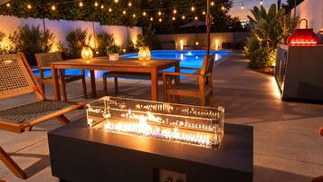 An outdoor patio with a glass-top fire feature, wooden furniture, string lights, and a pool in the background.
