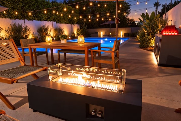 Eine Außenpatio mit einem Feuerstelle mit Glasplatte, Holzmöbeln, Lichterketten und einem Pool im Hintergrund.
