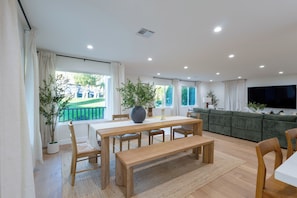 Ein geräumiges Wohnzimmer mit einem Holzesstisch, Bänken und einem grünen Sofa. Es gibt Blumentöpfe und einen großen Fernsehbildschirm.