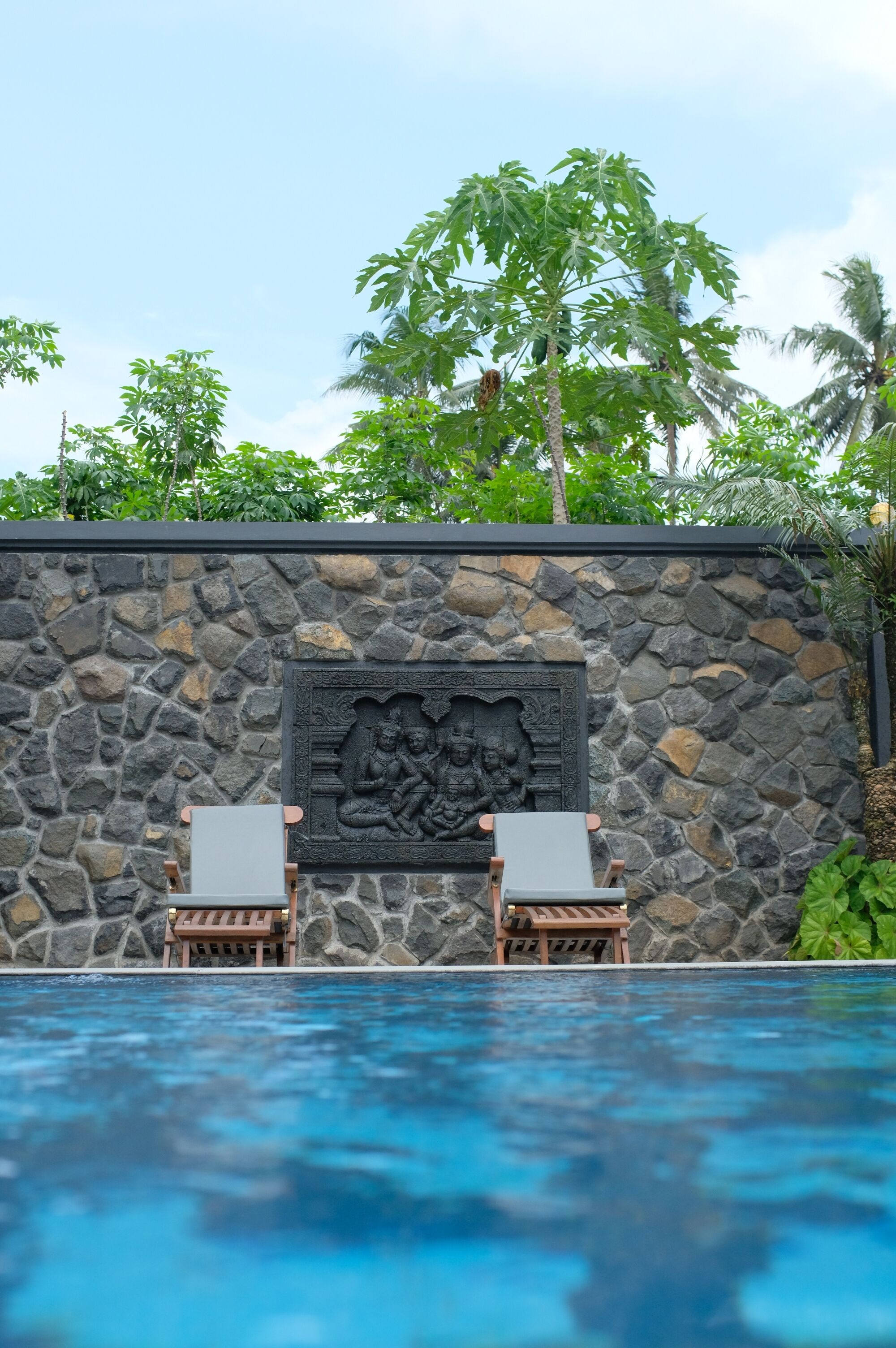 A stone wall with a relief sculpture, two lounge chairs, and a pool.