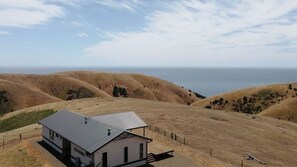 Une maison de plain-pied avec un toit en métal, entourée de collines couvertes d’herbe sèche et offrant une vue dégagée sur l’océan.