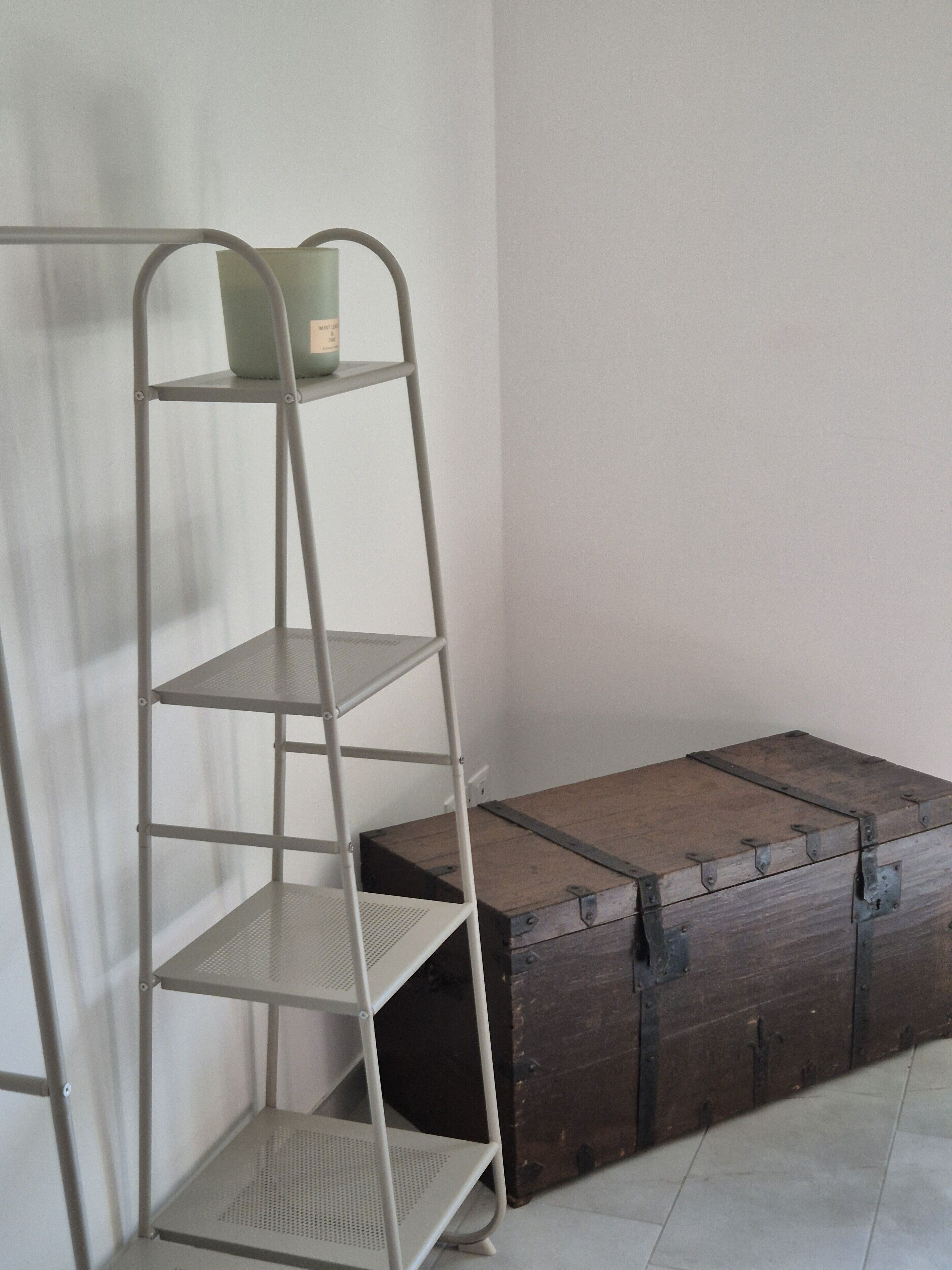 Una estantería metálica escalonada con un pequeño recipiente en la parte superior, junto a un cofre de madera con bandas metálicas.