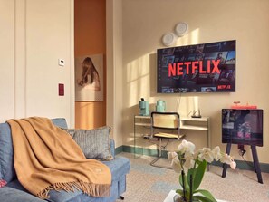 Un salon avec un canapé bleu, une table basse, un téléviseur affichant le logo de Netflix, un petit bureau avec une lampe et une étagère fixée au mur.