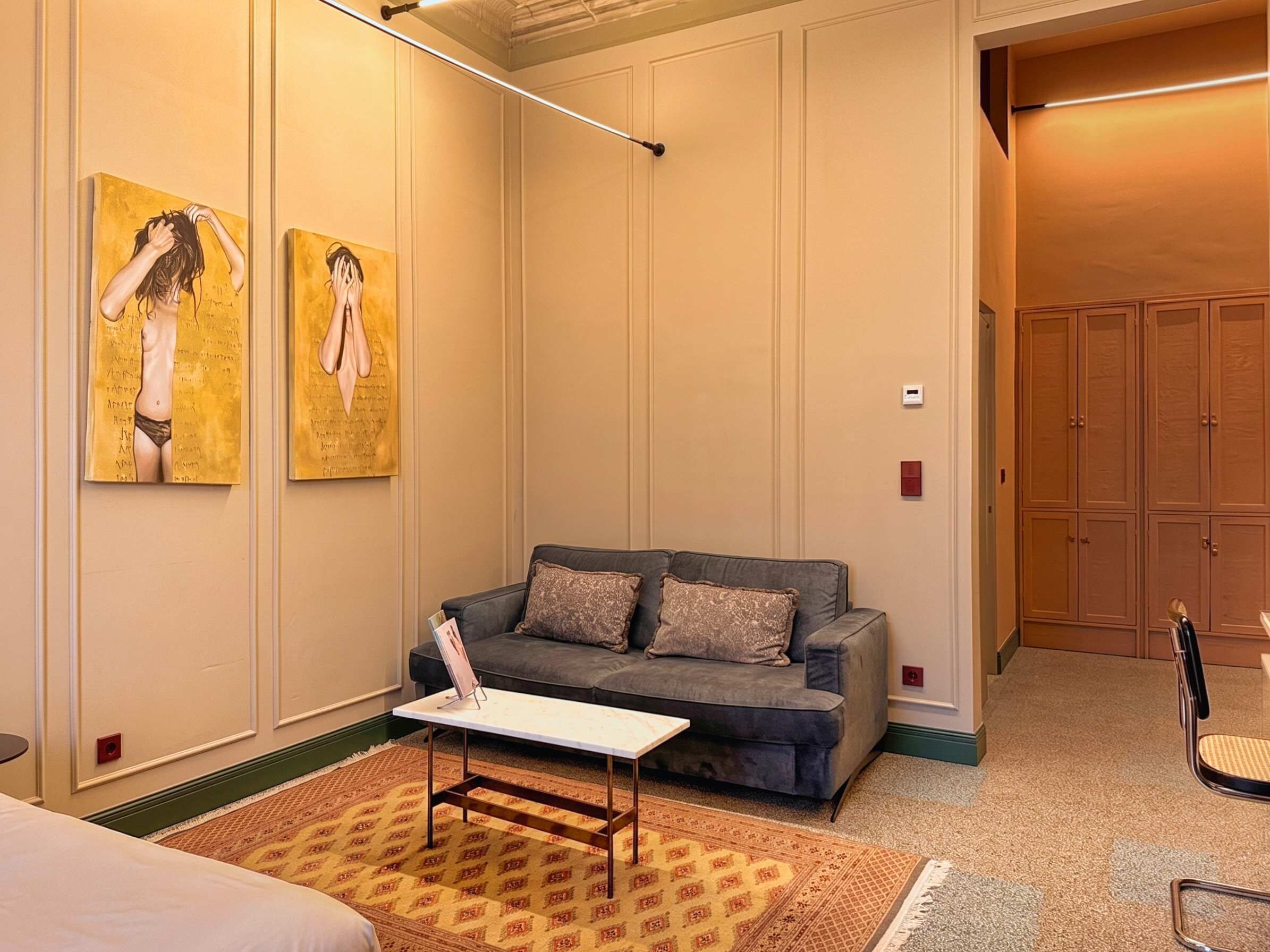 Habitación de hotel con un sofá gris, una mesita blanca pequeña, alfombras estampadas y dos cuadros en la pared.