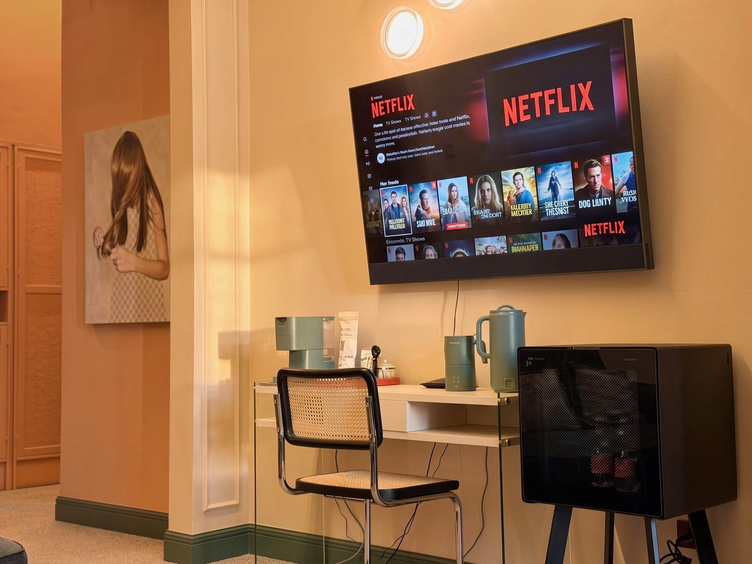 Une pièce équipée d’un téléviseur fixé au mur diffusant du contenu Netflix, d’un bureau avec une chaise, d’une machine à café et d’un écran d’ordinateur.