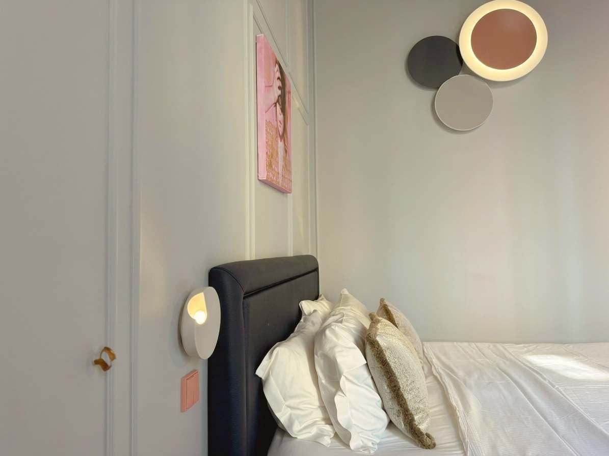 A bedroom with a bed, pillows, a wall-mounted light, and a circular mirror.