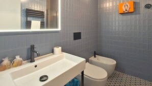 Ein Badezimmer mit einem weißen Waschbecken, einer Toilette und einem Spiegel. Auf dem Waschbecken liegen Pflegeprodukte und über dem Spiegel ein Handtuchhalter.
