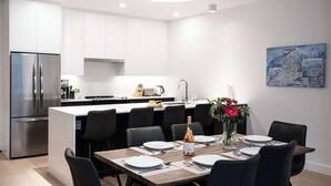 A modern kitchen with white cabinets, a stainless steel refrigerator, and a dining area with a wooden table set for four. - Revelstoke - Bluebird- New condo! (Revelstoke)