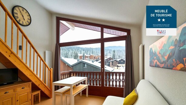 Una habitación con escalera de madera, un sofá blanco con almohada amarilla, una mesa de madera y un ventanal con vistas a edificios y árboles.