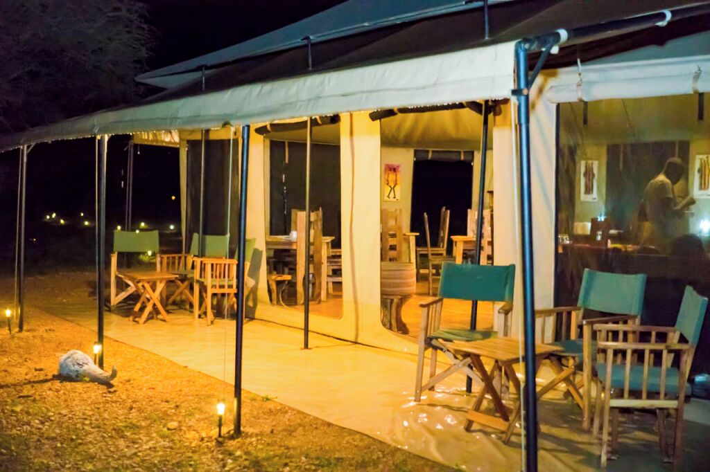 Un área de comedor al aire libre con carpas, mesas y sillas, iluminada por luces pequeñas.